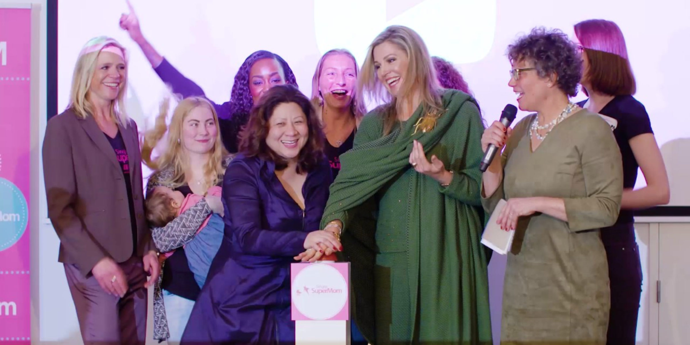 Group of women on a stage smiling and pushing a buzzer in front of a supermom backdrop