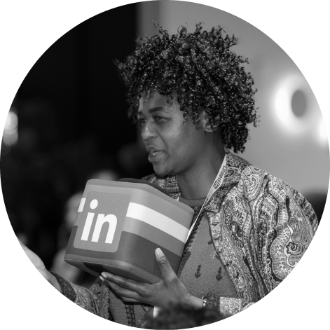 Woman with curly hair csmiling and carrying a box with the Linkedin logo on it. Photo in black and white.