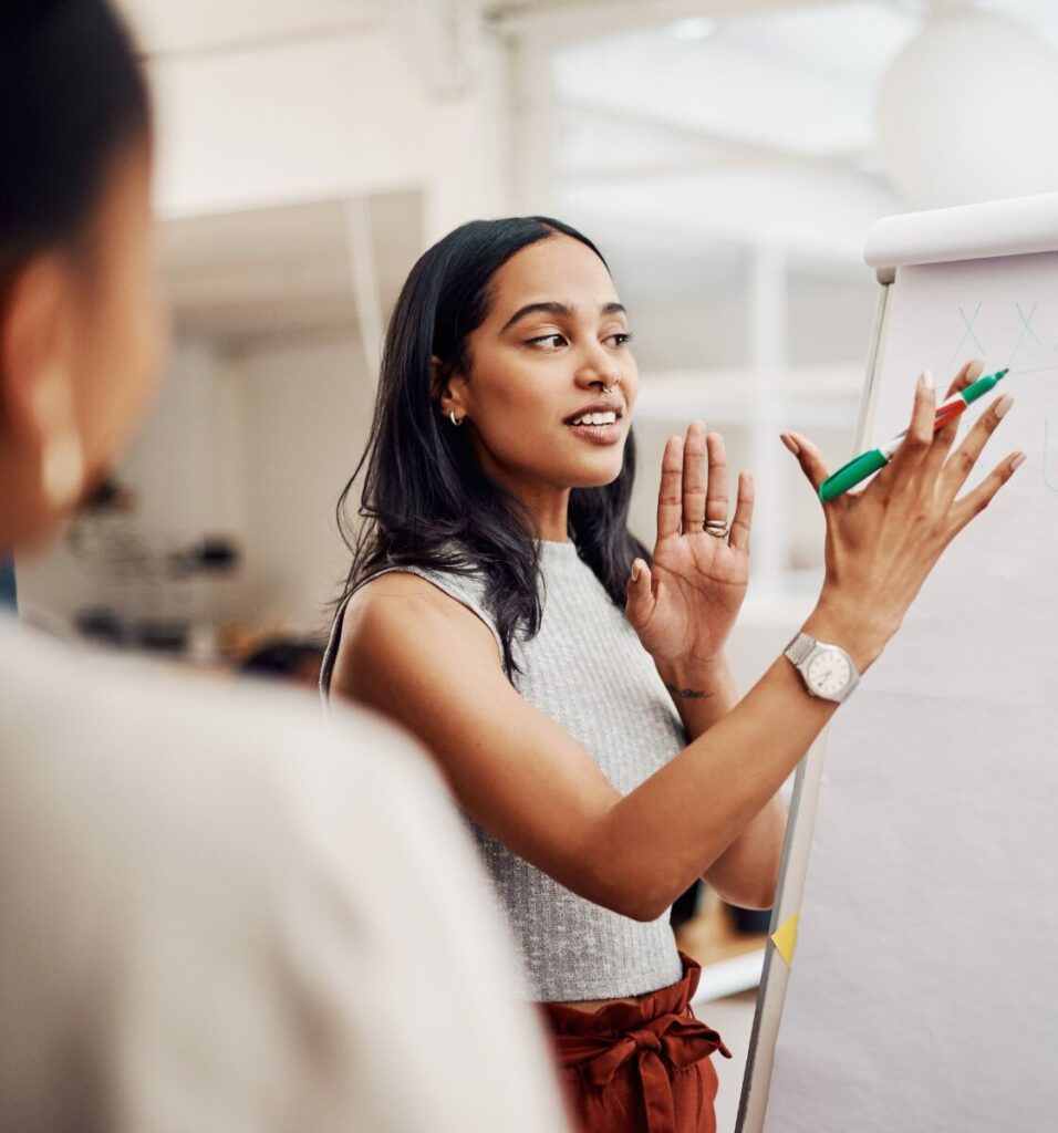 Young woman of colour writes on a flipchart while talking to an audience in front of her in a work environment.