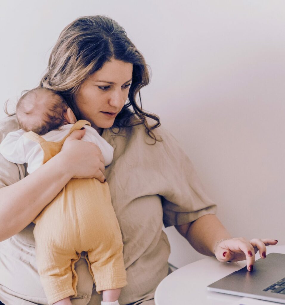 A person holding a baby while sitting at a desk with a laptop.