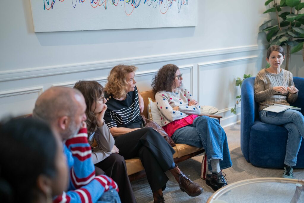 Five people sit on a sofa listening to a speaker sitting on an arm chair.